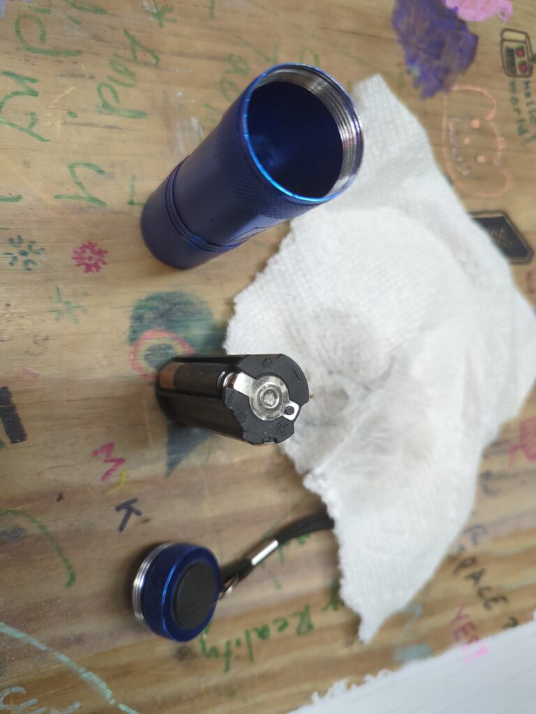 A flashlight, battery holster, and cap sit on the desk with an alcohol soaked paper towel in the backgrounu