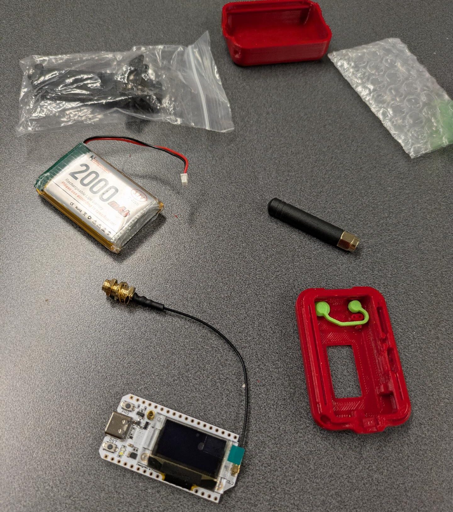 Parts of a Meshtastic lie on the table. It includes a circuit board containing the radio, a battery, an antenna, and a disassembled 3D printed case