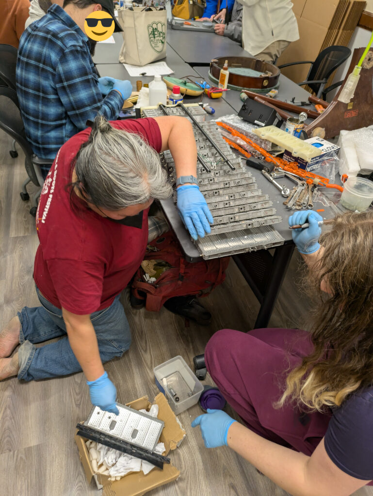 Prius battery cells separated and the owner is reaching for a cell on the ground. In the background, a lamp being repaired, and end table, and a table used with a walker.